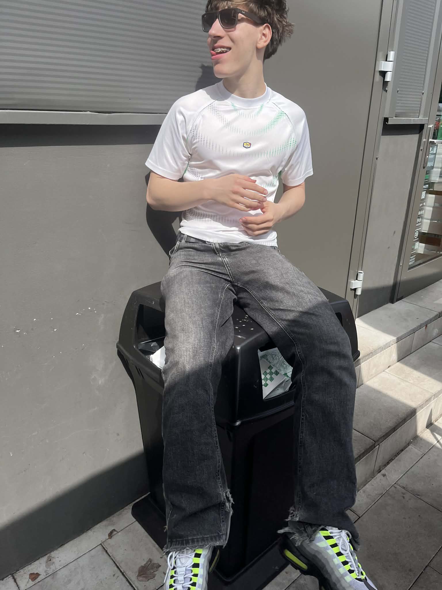 Fiatal férfi napszemüvegben, fehér streetwear pólóban, szürke farmerben és sportcipőben ül kint