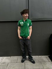 Fiatal férfi zöld Ralph Lauren pólóban, fekete farmerben, utcai stílusban, szürke fal előtt.